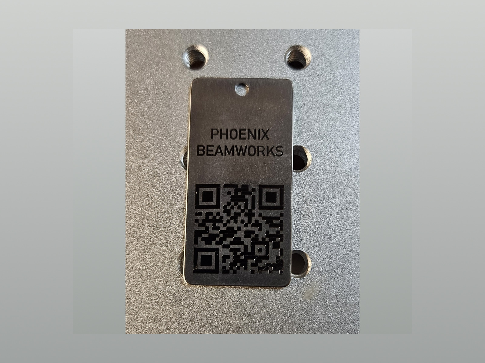 Bare stainless steel Phoenix Beamworks key or ID tag with black annealed lettering and QR code on a metal shop fixture