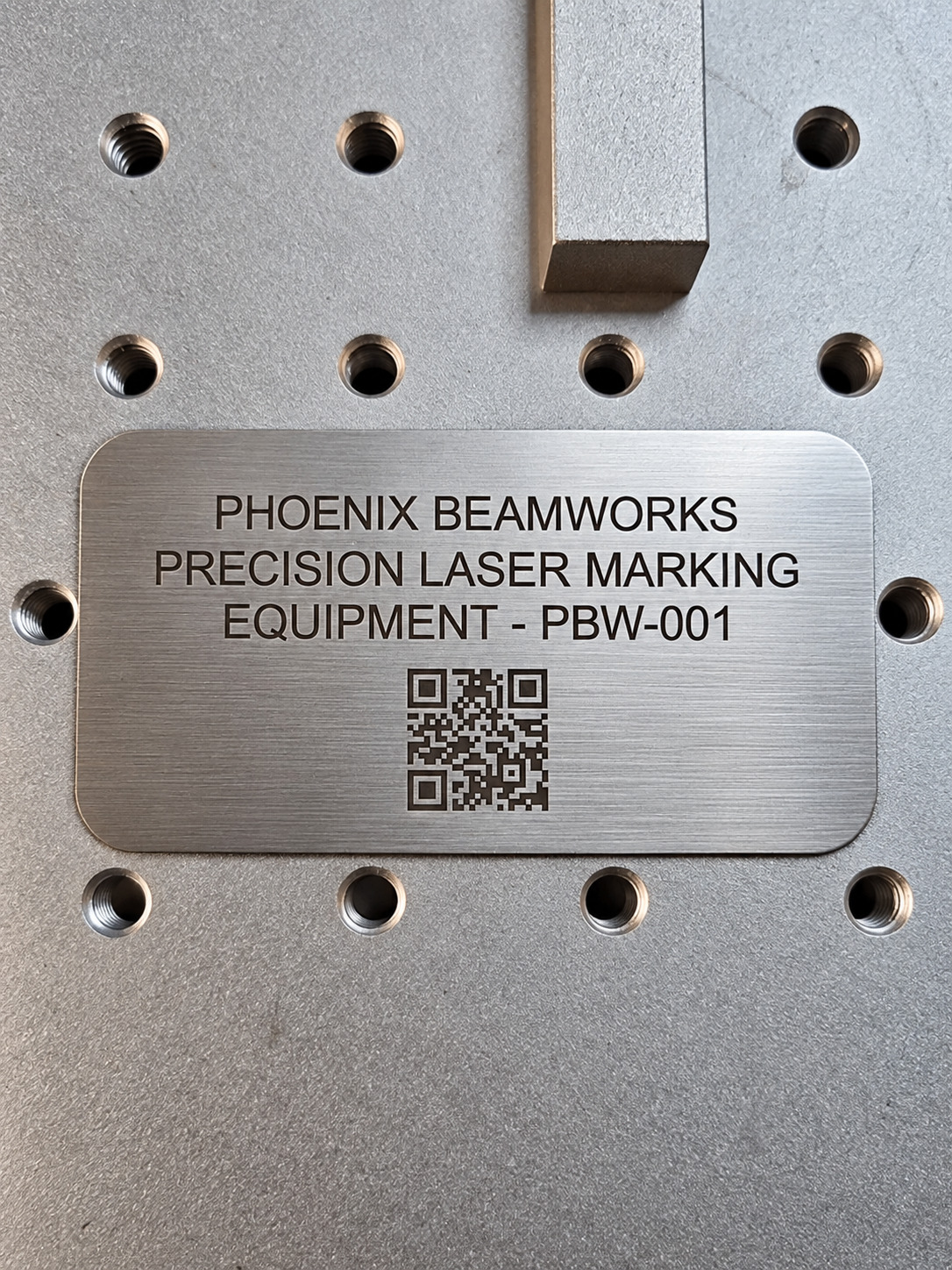 Brushed stainless Phoenix Beamworks equipment plate with engraved serial number and QR code on the laser worktable